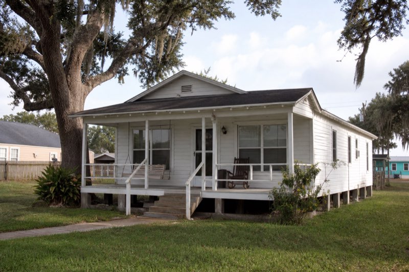 Mid-century white pier-and-beam cottage under mature oaks in a Texas bay town