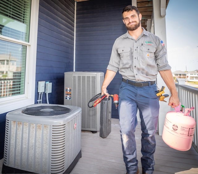 Mackey Services technician carrying tools beside two outdoor AC condenser units